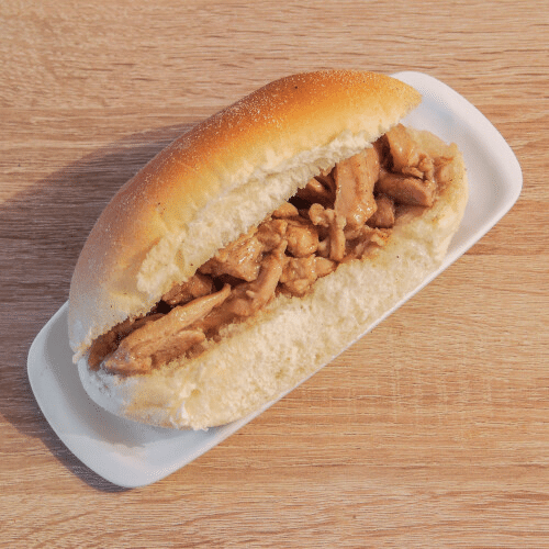 Een broodje getrokken kip ligt op een wit bord op een houten tafel.