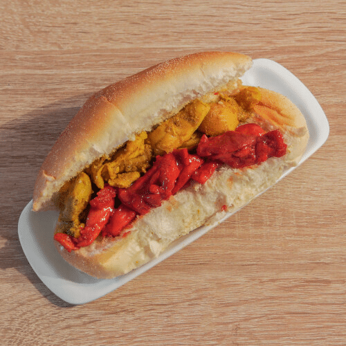 Een hotdog met kip en rode paprika op een broodje op een witte plaat op een houten tafel.