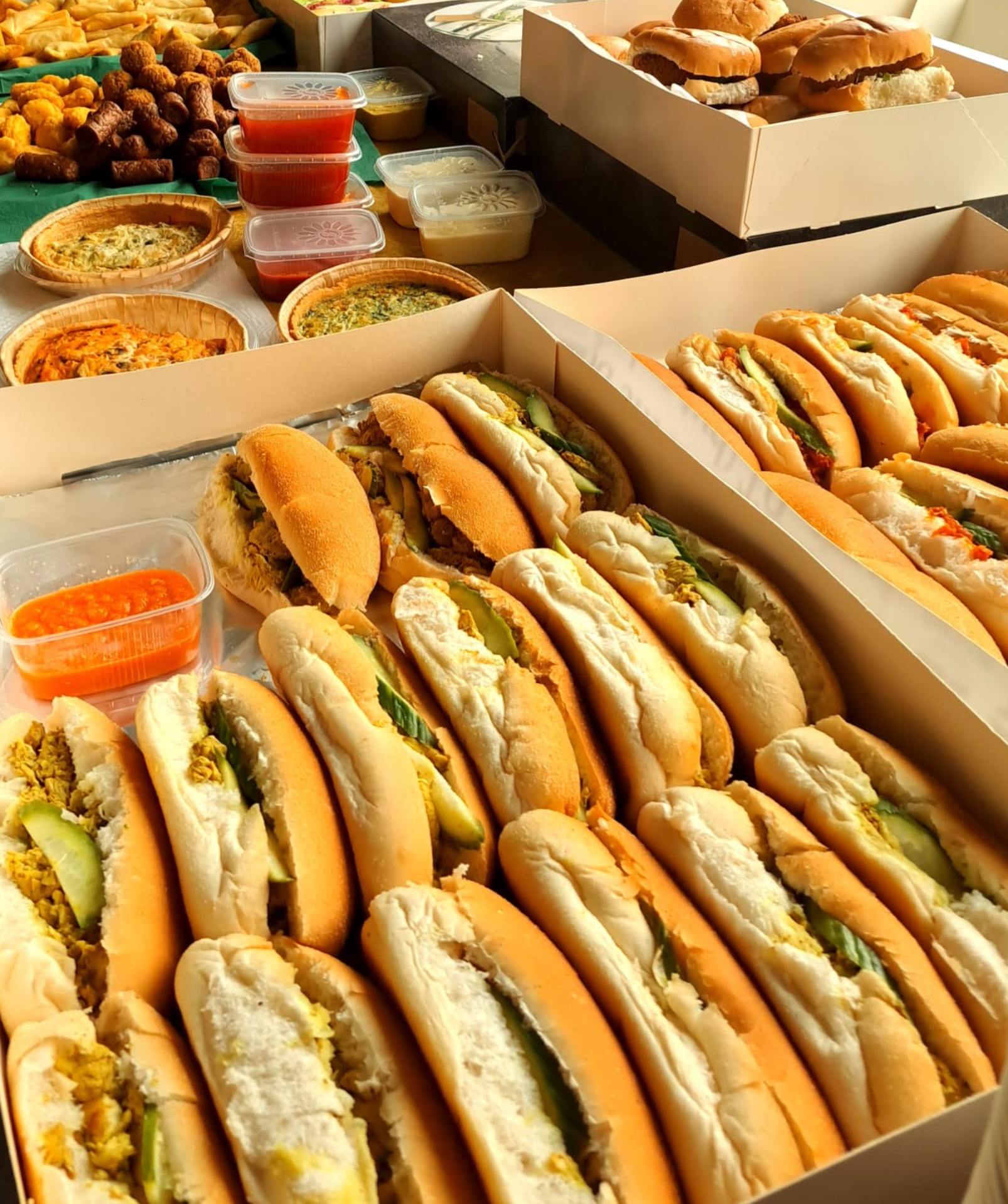 Close-up van een tafel met dozen hotdogs, hamburgers, taarten en kleine bakjes sauzen.