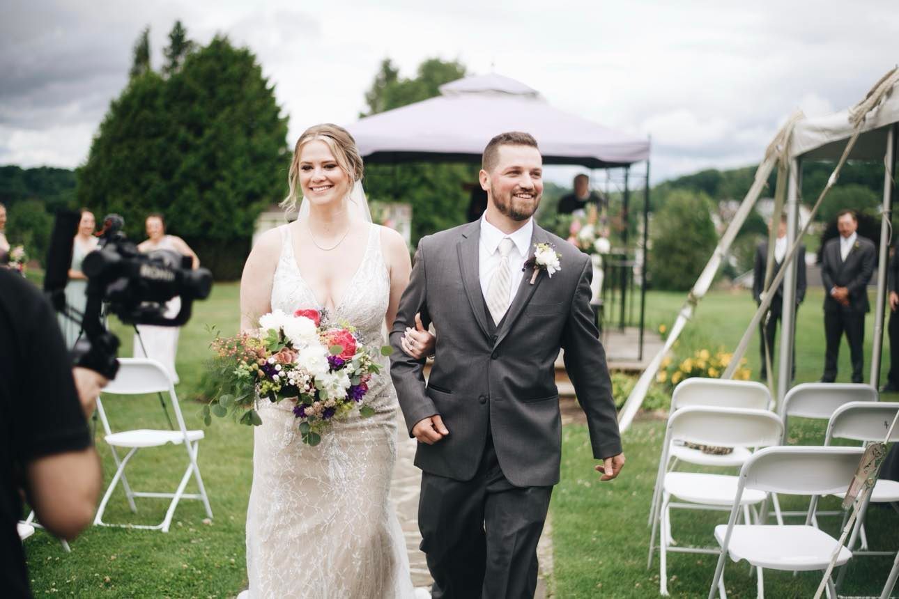 A smiling bride and groom walk hand-in-hand down an aisle at an outdoor wedding ceremony, with guests and chairs nearby.