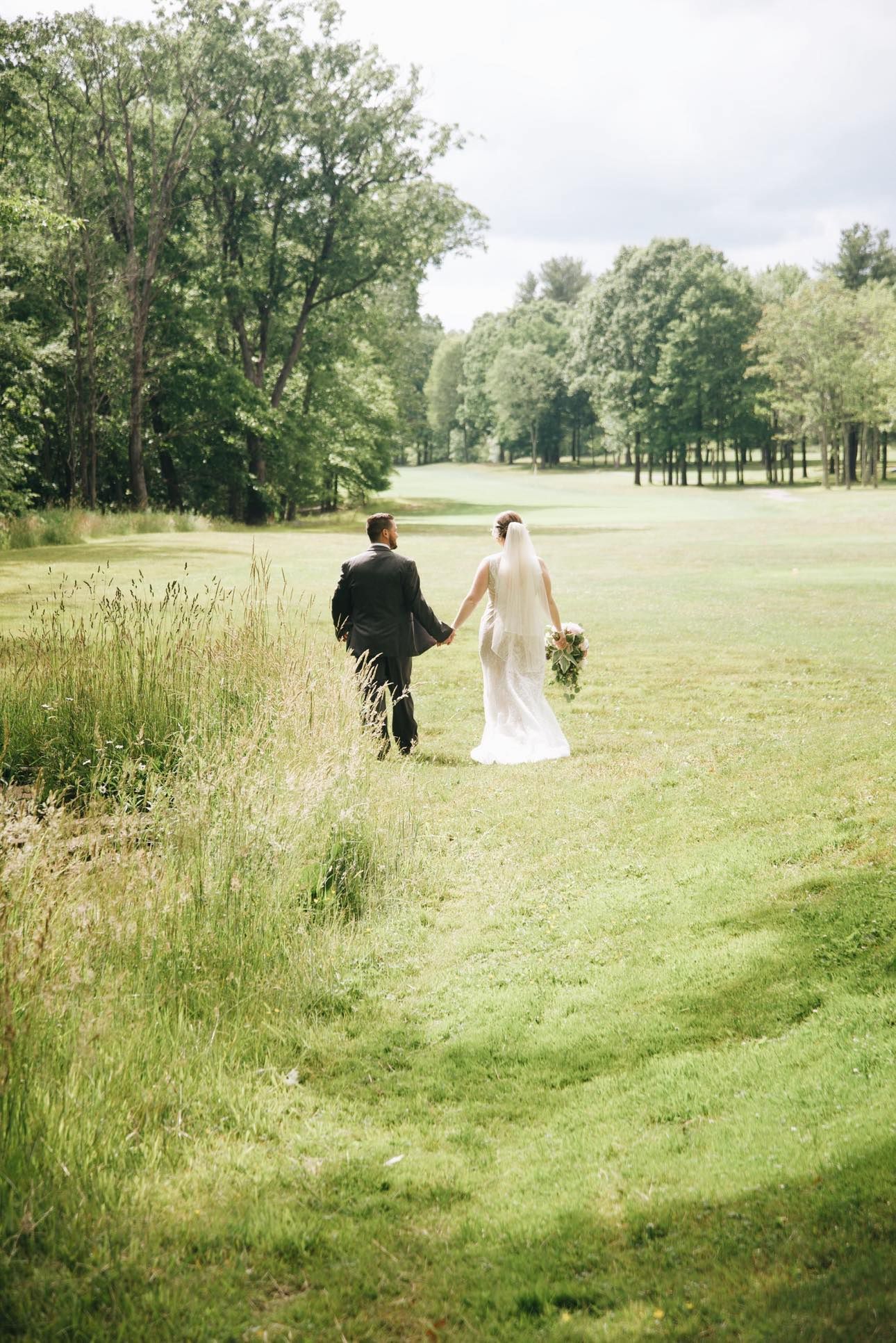 A bride and groom walk hand-in-hand through a grassy meadow toward a line of trees on a bright day.