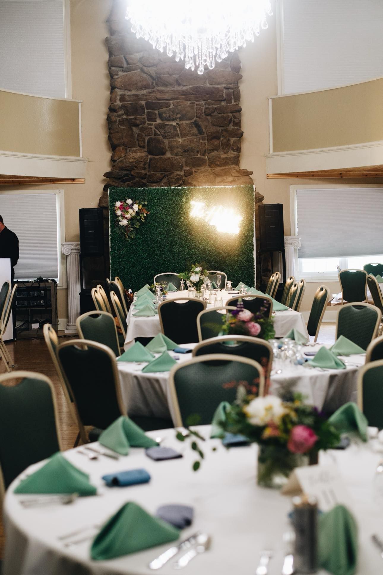 A banquet hall with round tables set for an event, featuring green napkins, floral centerpieces, and a moss-covered wall.