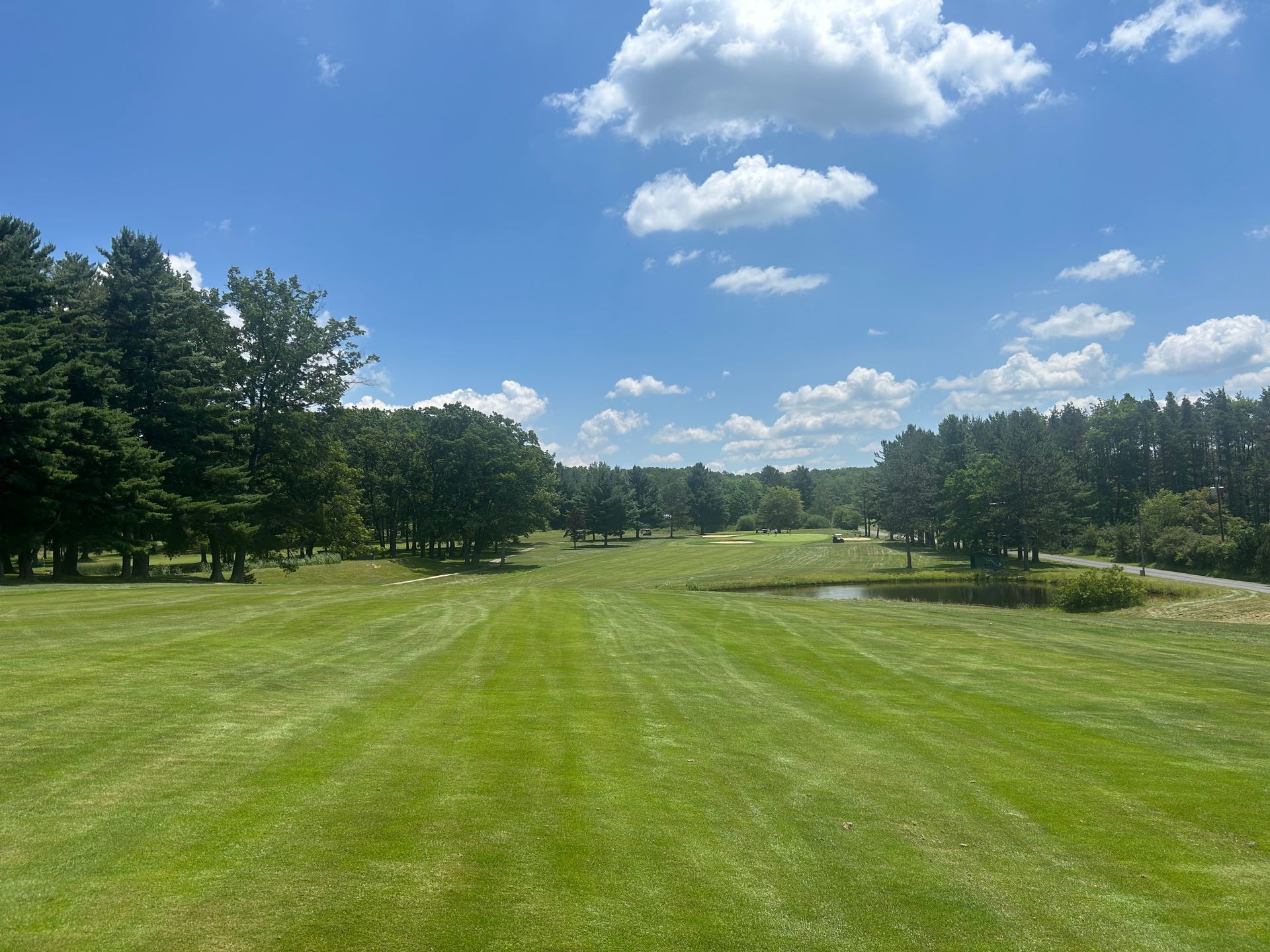 Green golf course under a bright blue sky with scattered clouds. Trees line the sides of the fairway.