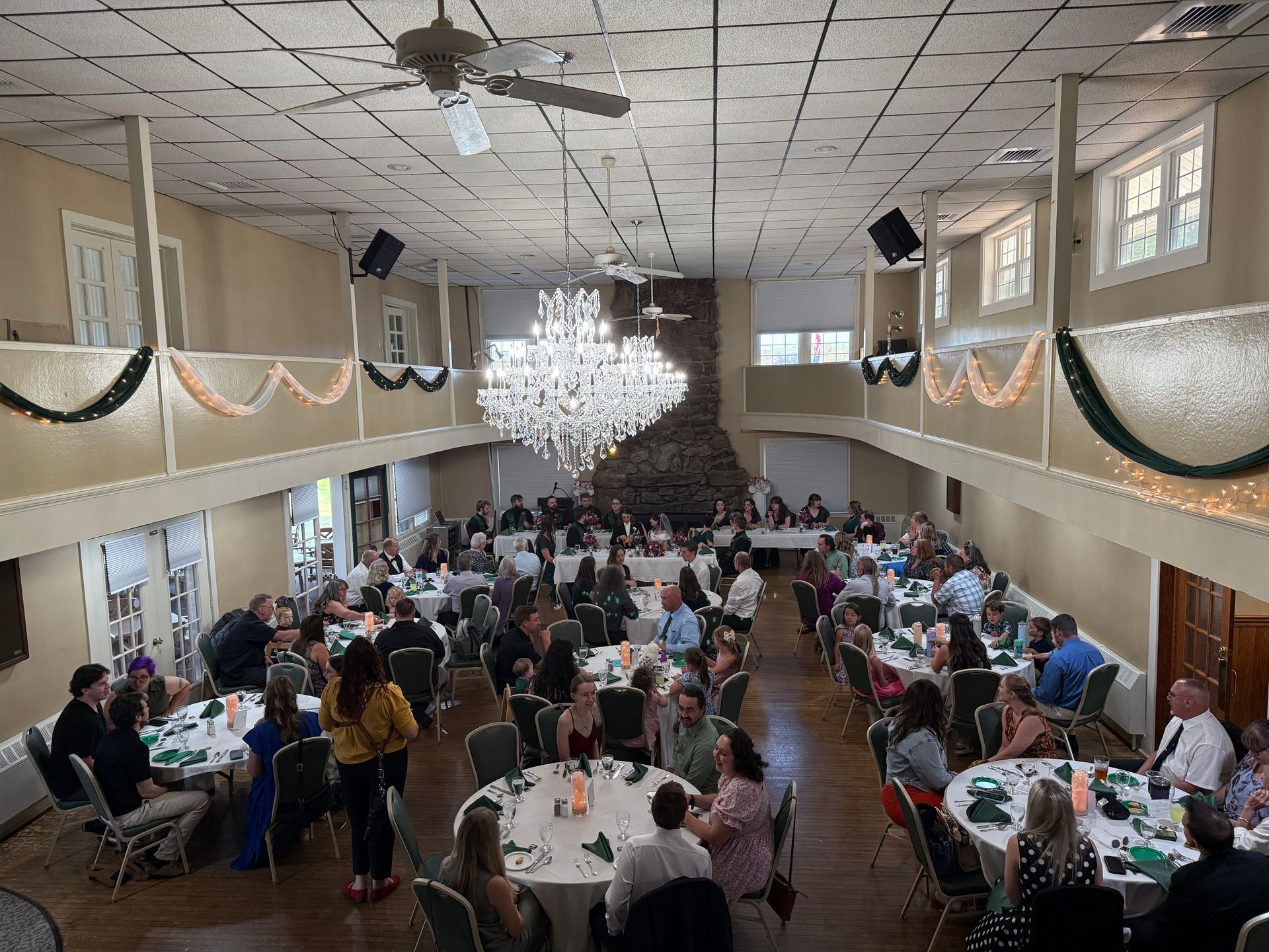 A banquet hall with a large crystal chandelier, tables occupied by guests, and draped garlands along the upper balcony.