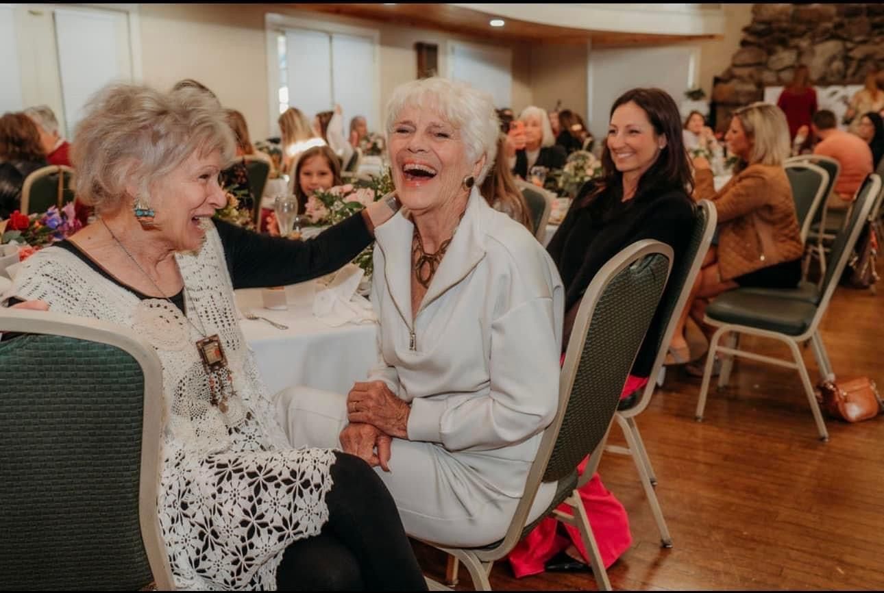 Two people laughing while sitting at a table in a brightly lit event room filled with other guests.