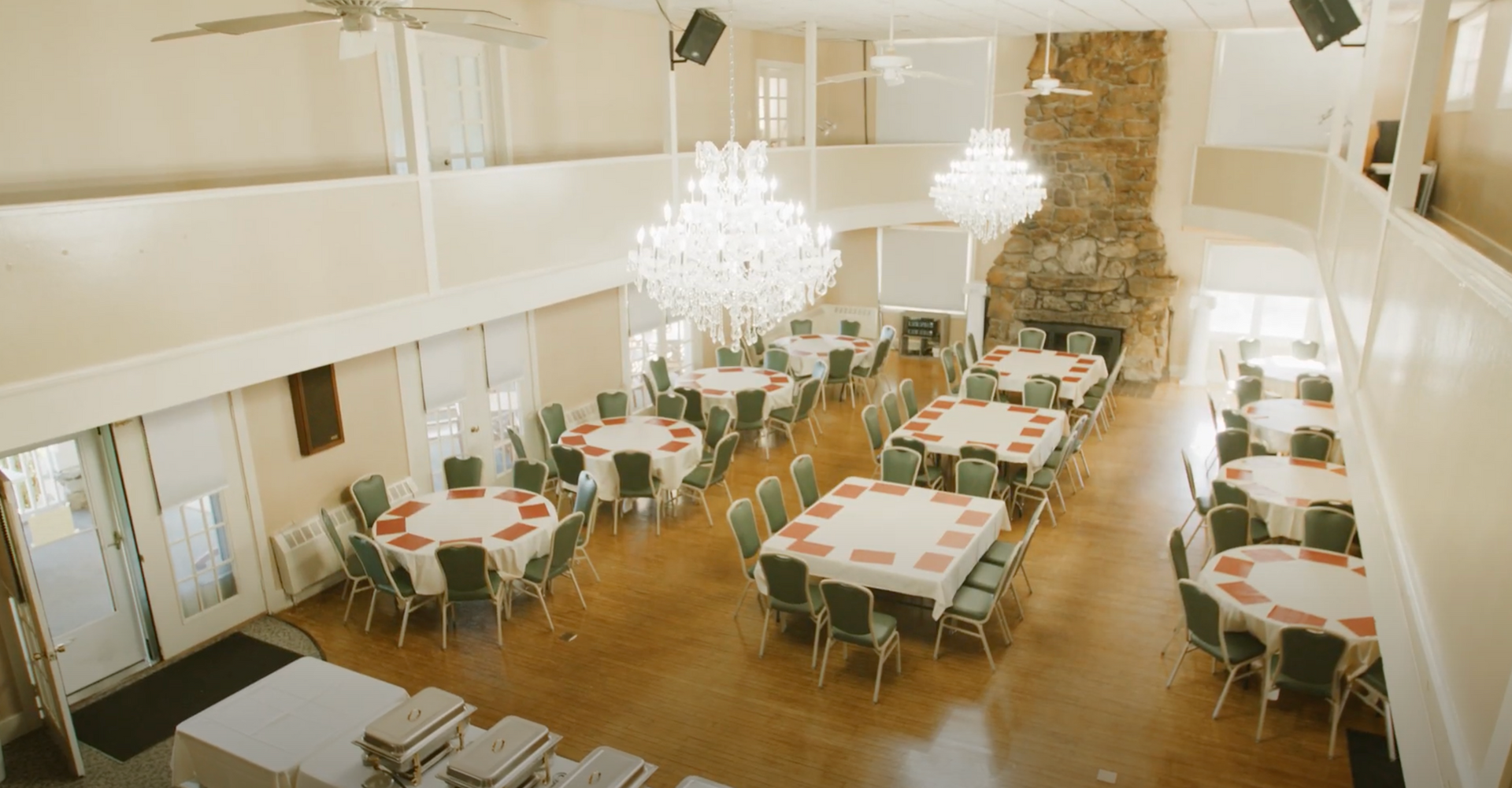 High-angle view of a banquet hall with round and square tables, chandeliers, a stone fireplace, and light-toned decor.
