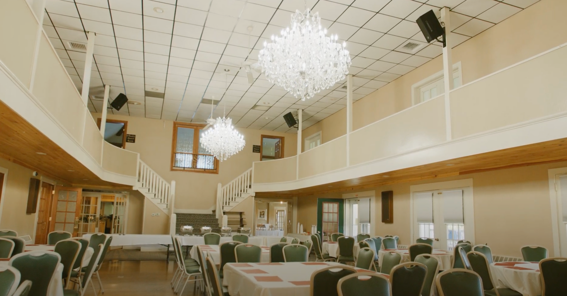 A spacious event hall with two large crystal chandeliers, a balcony level, and tables set with chairs for a gathering.