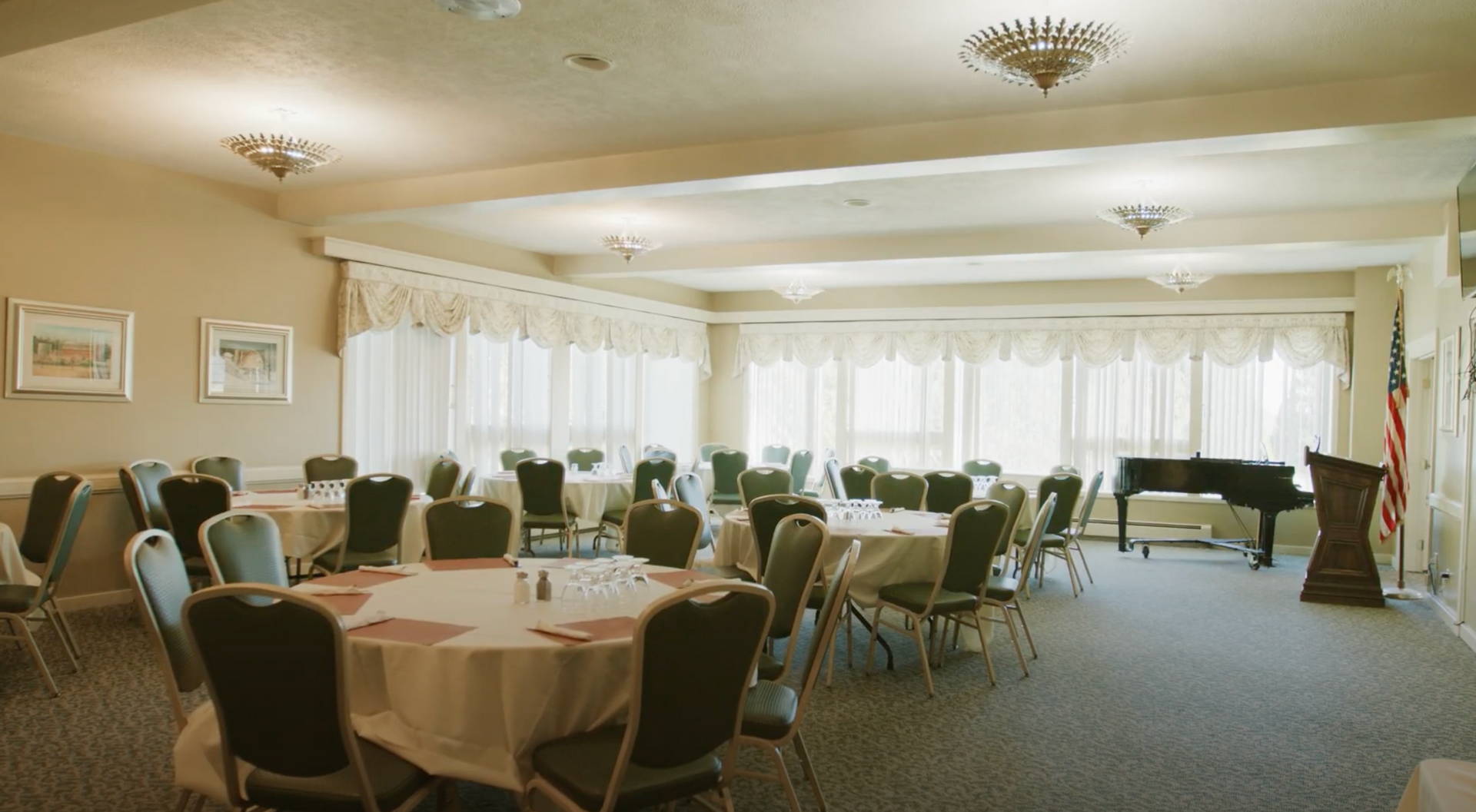 A bright, carpeted banquet hall featuring several round tables with white tablecloths, dark chairs, and a grand piano.