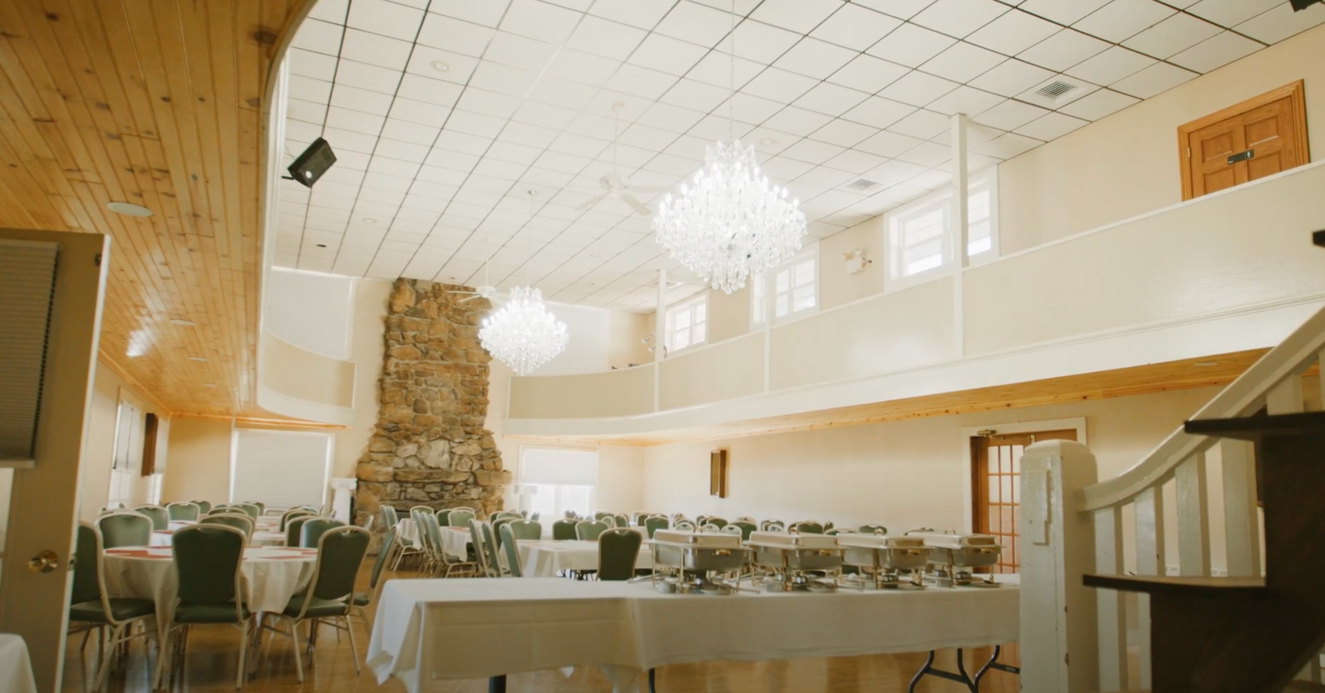 A banquet hall with tables, green chairs, two crystal chandeliers, and a large stone fireplace under a high ceiling.