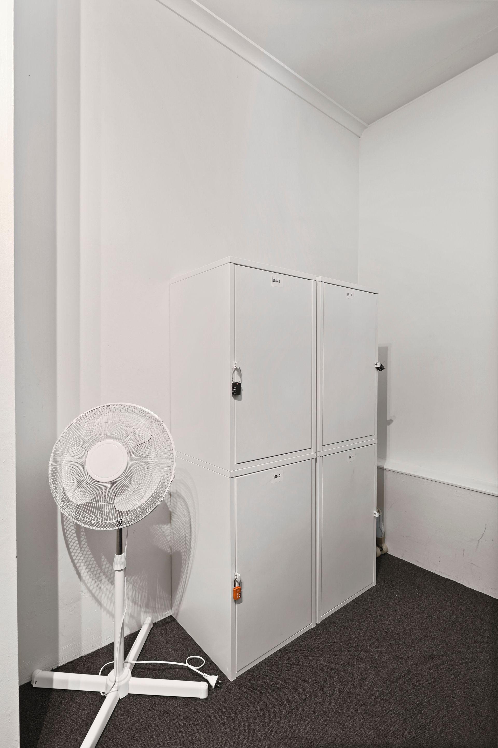 A white fan is sitting in the corner of a room next to lockers.