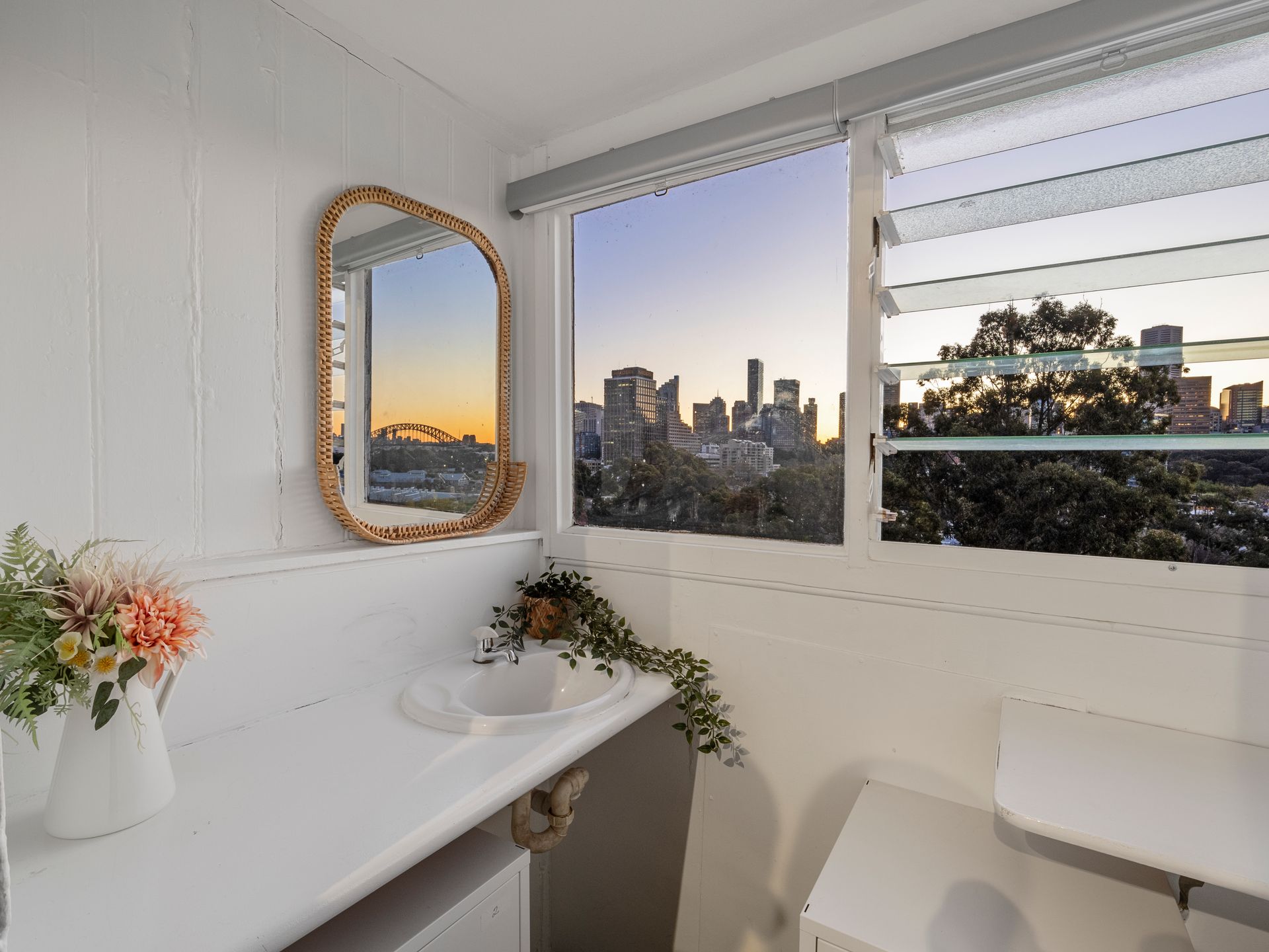 A bathroom with a sink , toilet , mirror and window with a view of the city.