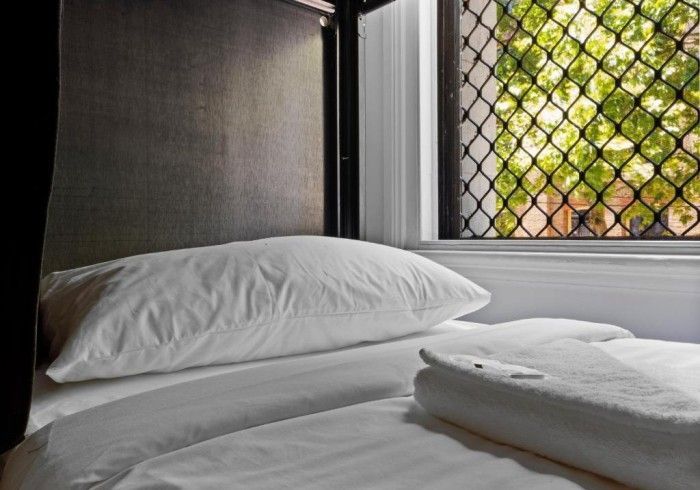 A bed with white sheets and pillows next to a window with a chain link fence in the background.