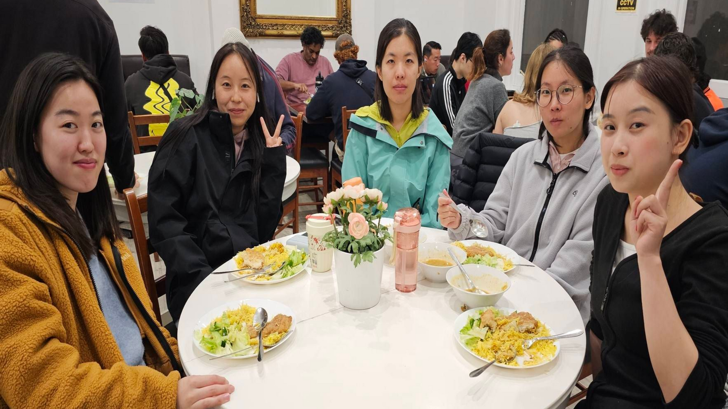 Two young women with curly hair seated at a table with food. One smiles and wears glasses, the other gives a thumbs up.