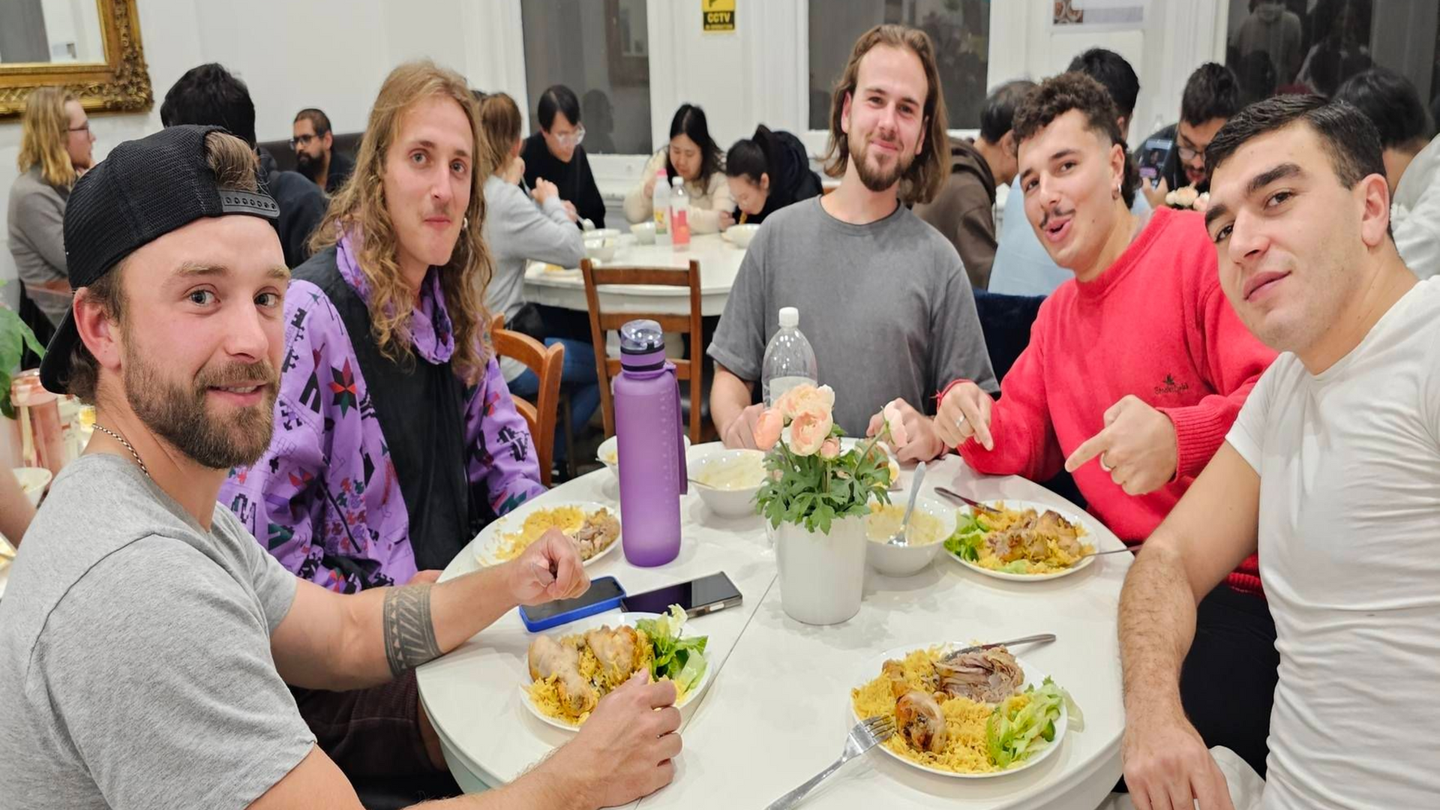 Five young adults at a round table eating a meal in a dining hall.