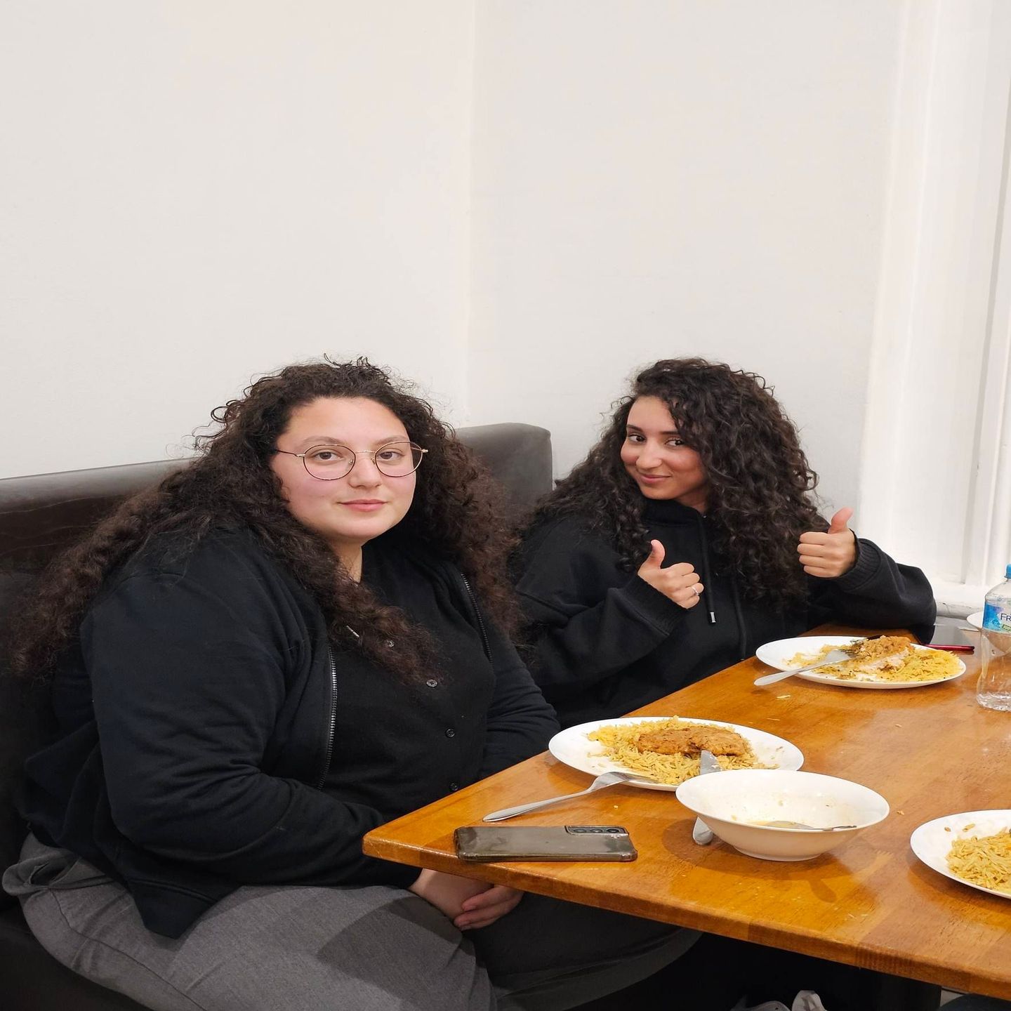 Two young women with curly hair seated at a table with food. One smiles and wears glasses, the other gives a thumbs up.