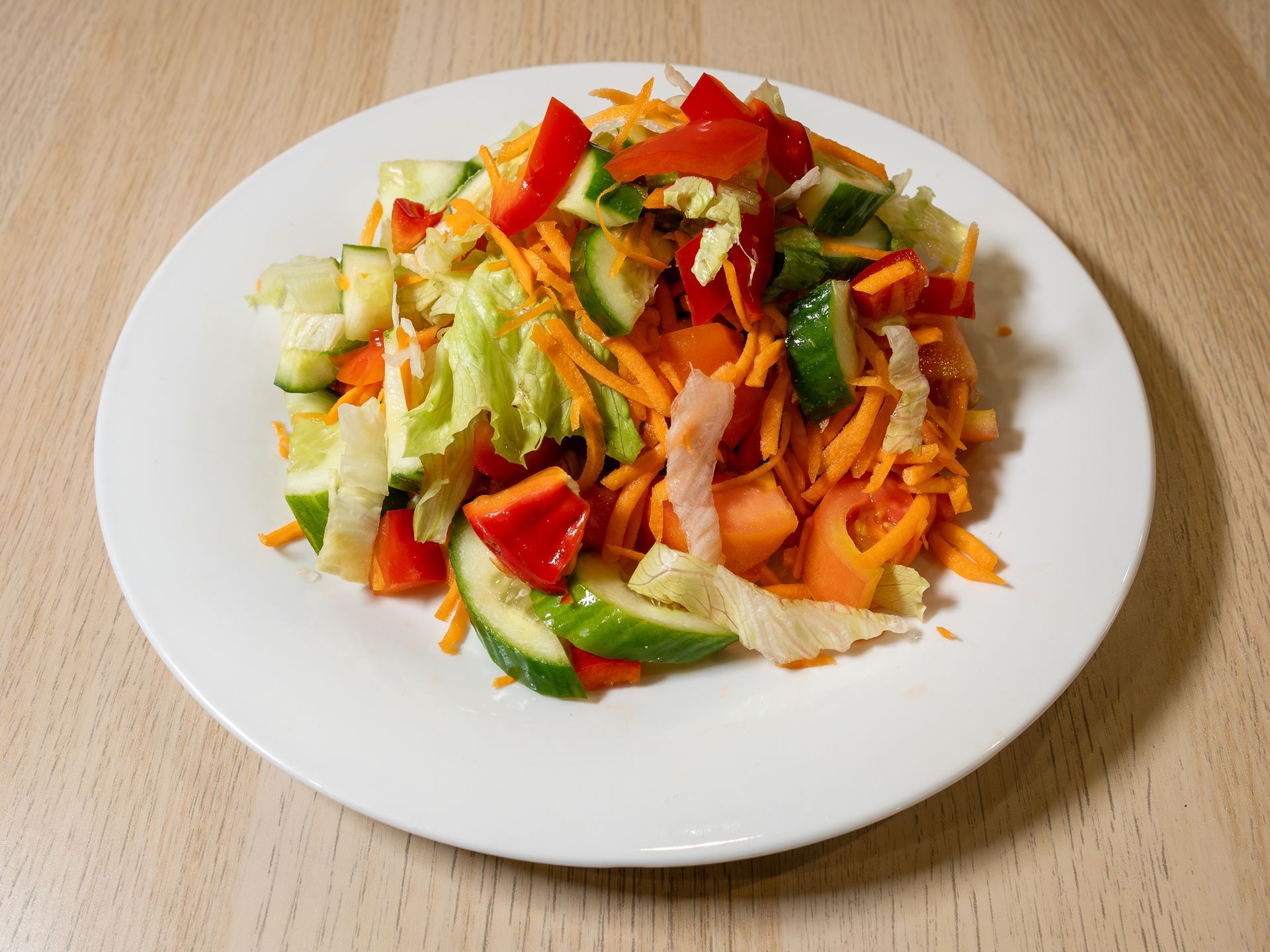 A white plate topped with a salad of carrots , cucumbers , peppers and lettuce on a wooden table.