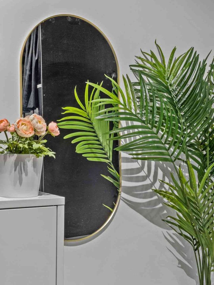 Oval black mirror with gold trim hanging on a gray wall, next to a potted plant and faux palm leaves.
