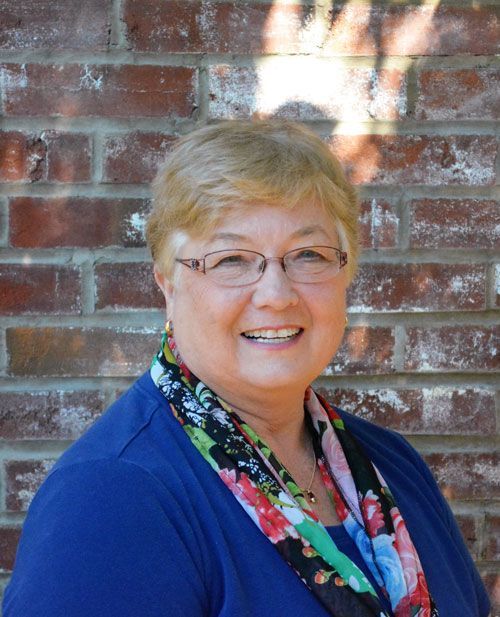 A woman wearing glasses and a scarf is smiling in front of a brick wall