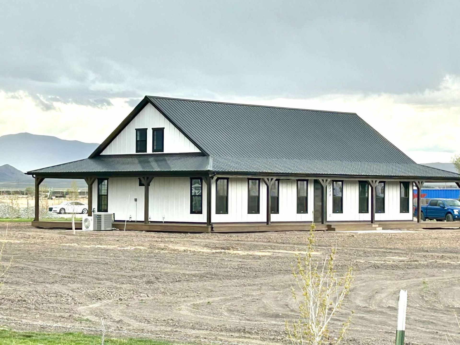 A large white barndominium  with a black roof is sitting in the middle of a dirt field.