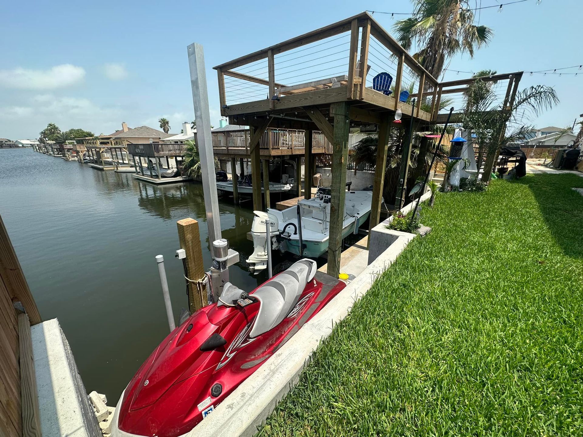 Jet ski and boat dock on a canal with a multi-level wooden structure, green grass, and houses.
