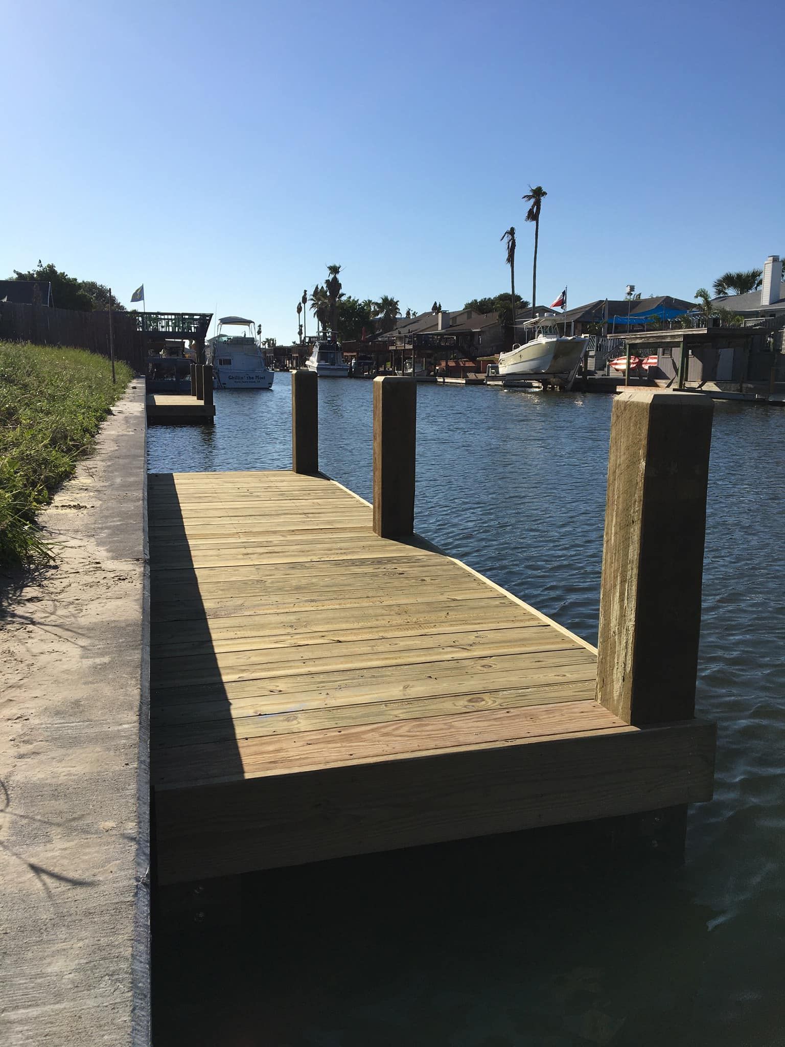 Wooden dock extending into a canal, with boats and buildings visible in the background under a blue sky.