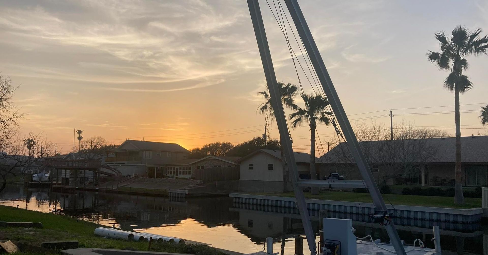 Sunset over a canal with houses and palm trees, power lines in the foreground, water reflecting the sky.