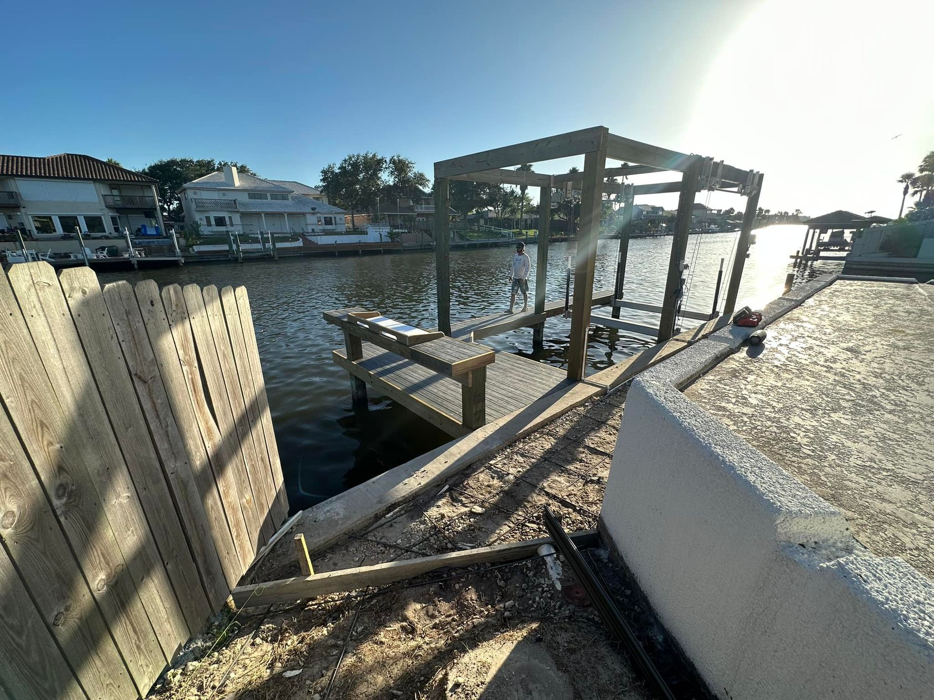 Dock under construction on a canal, with a wooden framework and surrounding houses in the background. Bright sunlight.