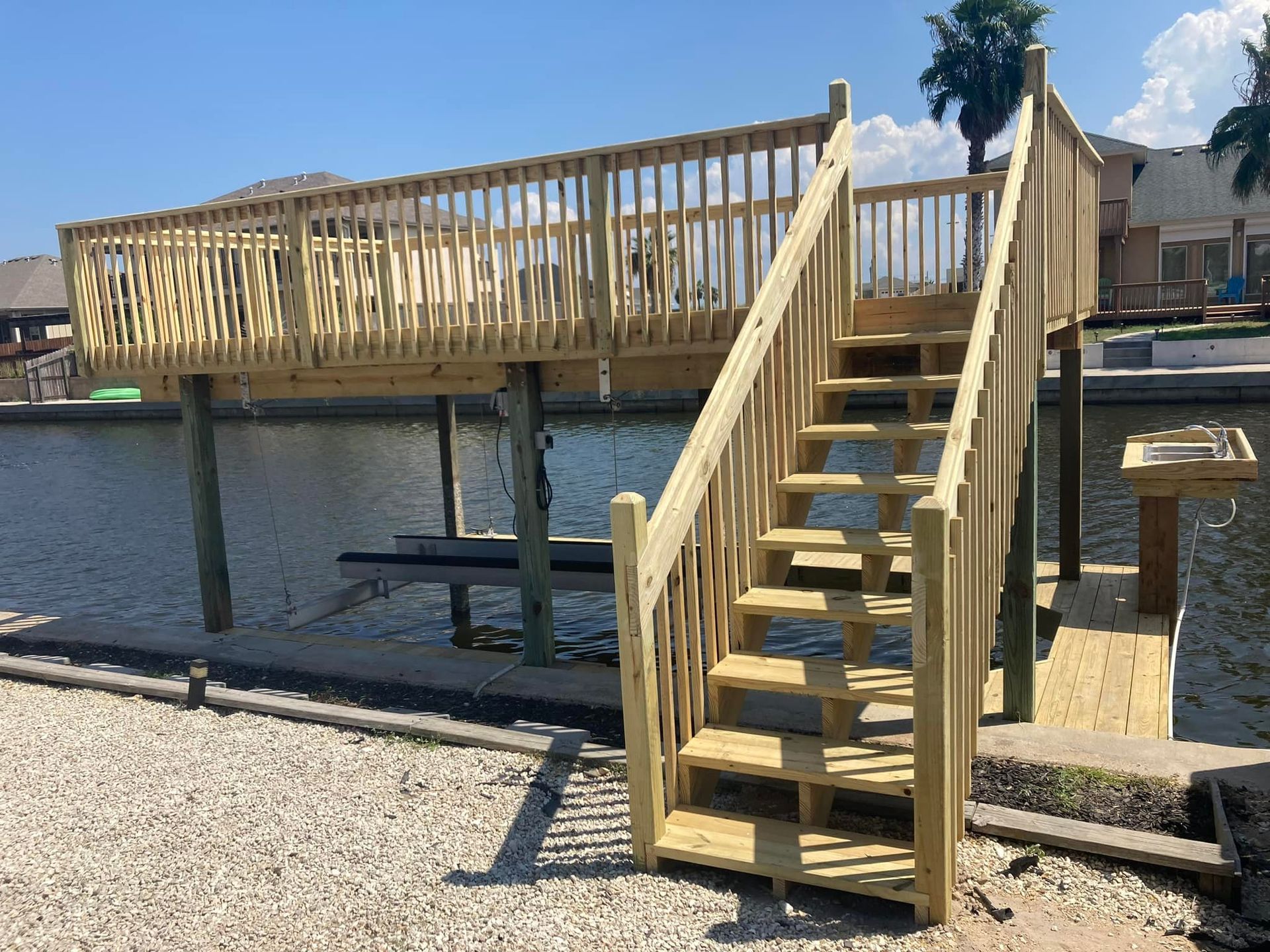 Wooden deck with stairs over water, railings, posts, and gravel ground.