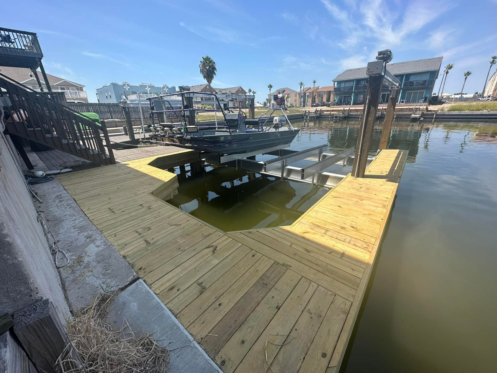 Wooden dock with boat lift over water, houses in background, sunny day.