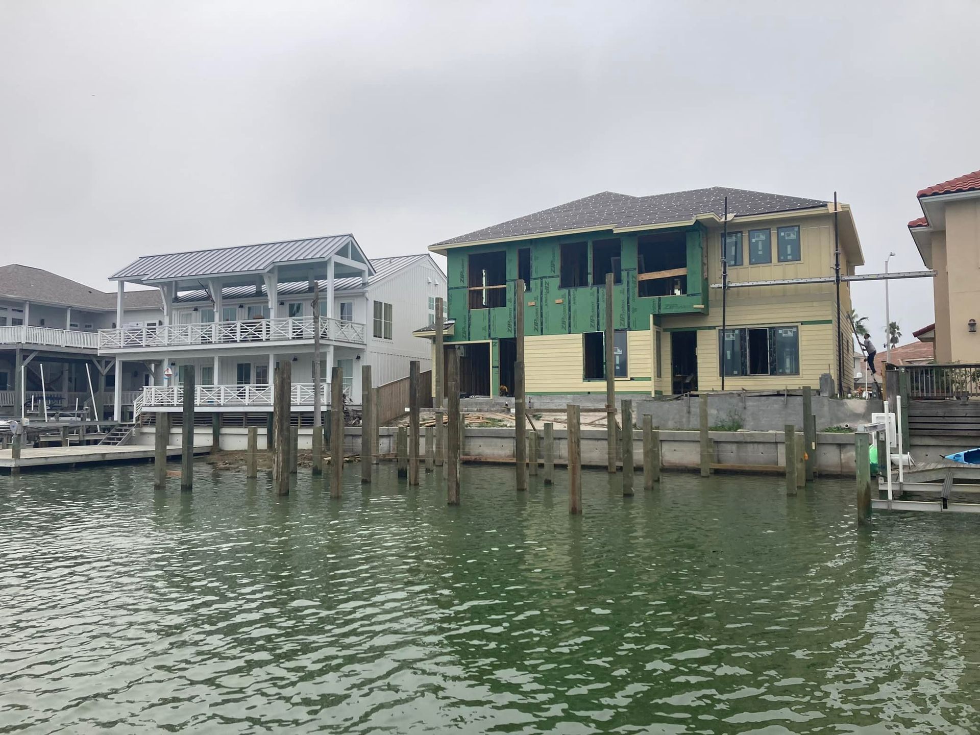 Houses under construction along a waterway, supported by wooden pilings. Overcast sky.