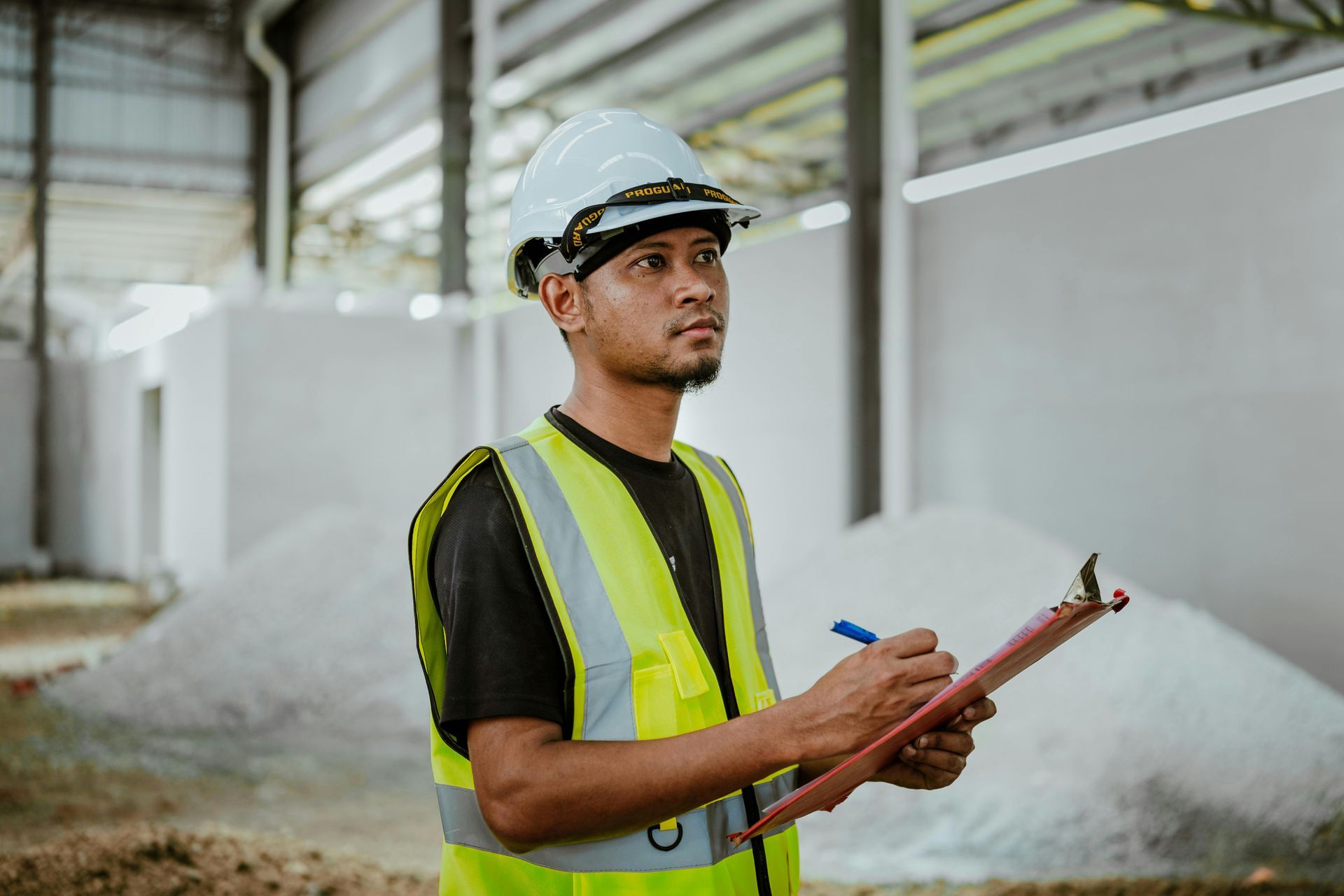 Man in hard hat and vest, holding clipboard, surveys a warehouse.