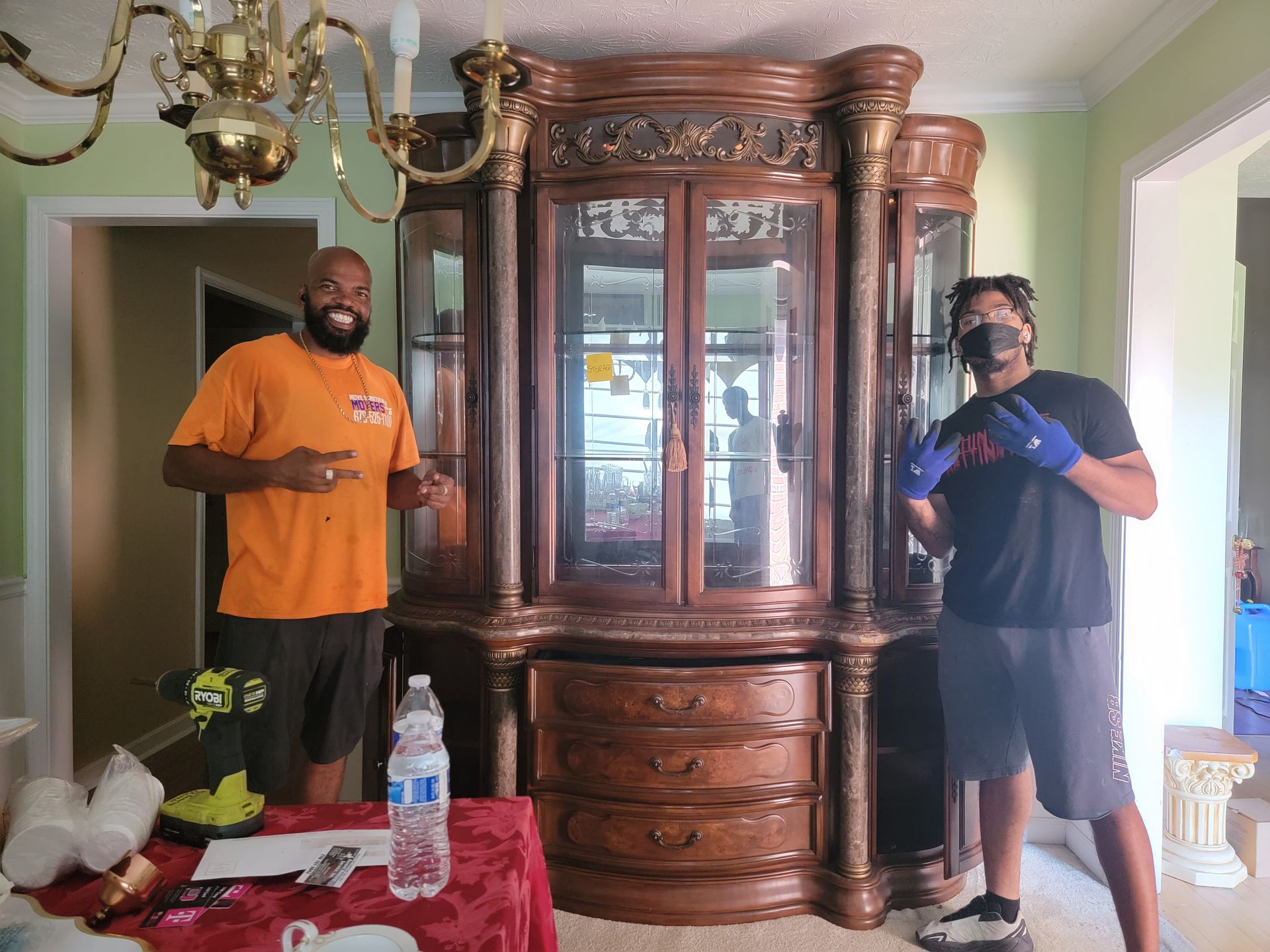 Two men with a large, ornate cabinet. One points at it and smiles. The other wears gloves and makes a peace sign.