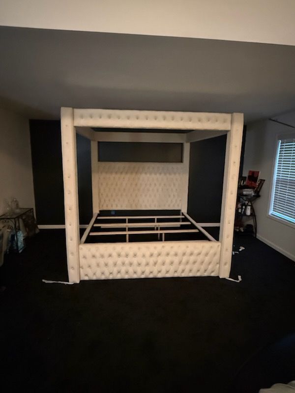 White upholstered canopy bed frame on black carpet in a room.