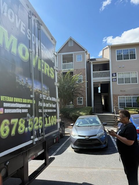 Moving truck parked outside an apartment building. A person stands nearby, holding a phone.