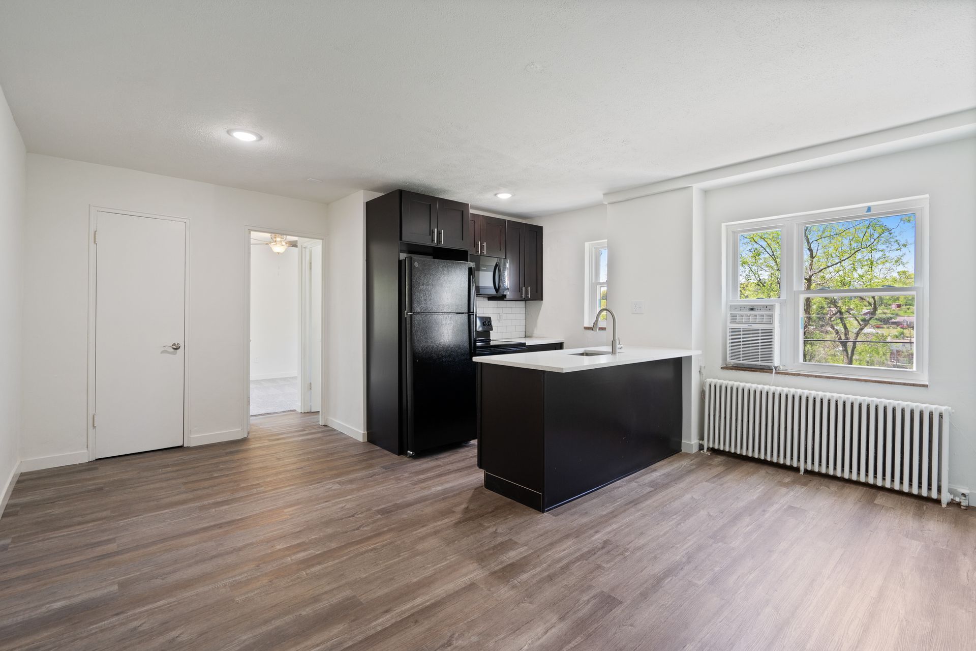 An empty living room with a kitchen and a refrigerator.