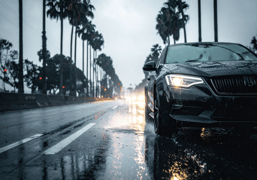 Car driving on a rain-soaked city street with headlights on and reflections on wet pavement.