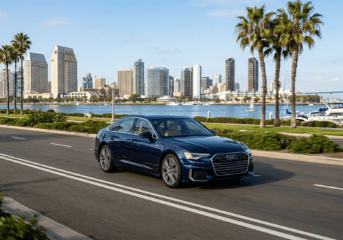 Blue Audi sedan driving along a waterfront city boulevard with palm trees, marina views.