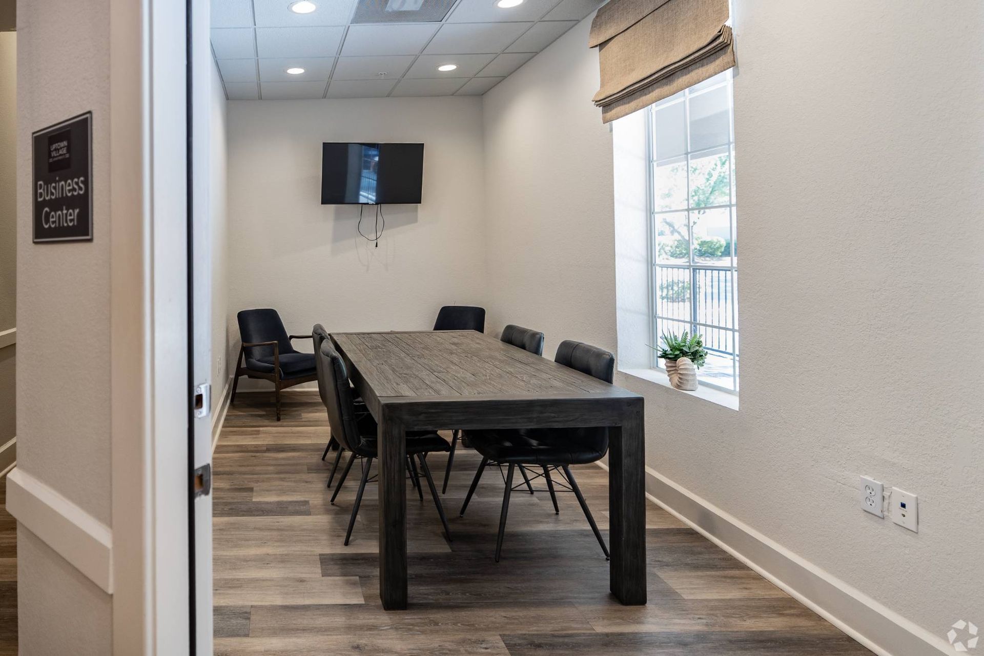 Community business center with a large wooden conference table, black chairs, wall-mounted TV, and a window.