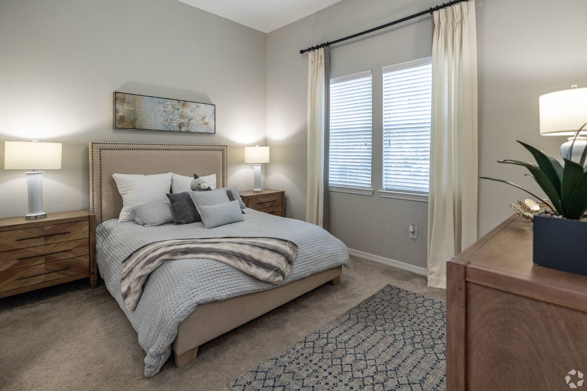 Bedroom in an apartment with a beige upholstered bed, two nightstands, lamps, and window with blinds
