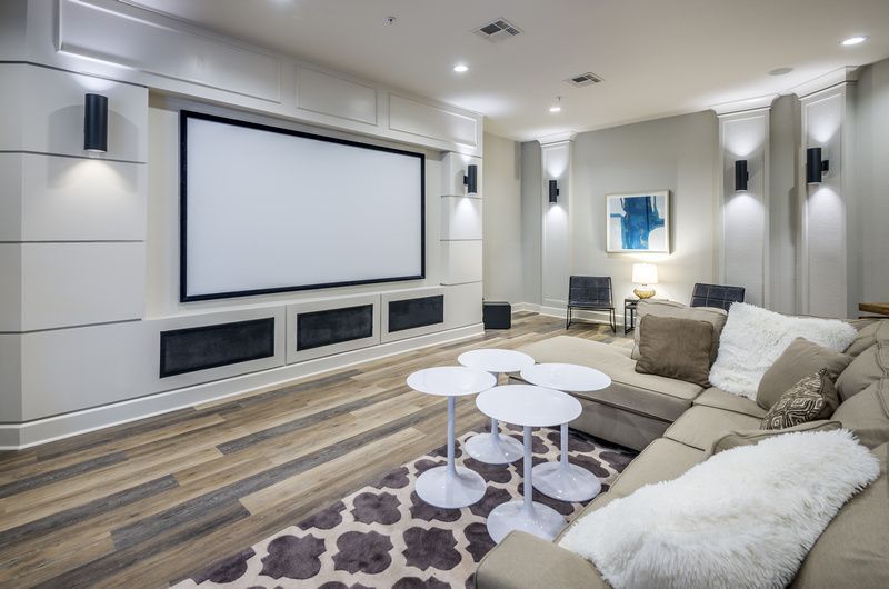 Modern community lounge with a large wall-mounted screen, beige sectional sofa, and white round tables.