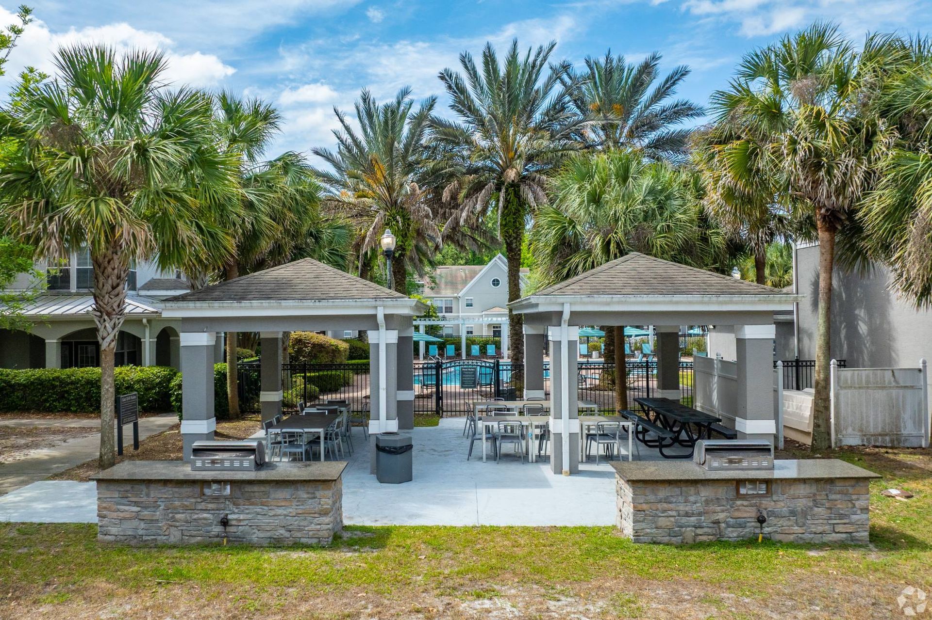 Outdoor community pool area with palm trees, shaded pavilions, and seating.
