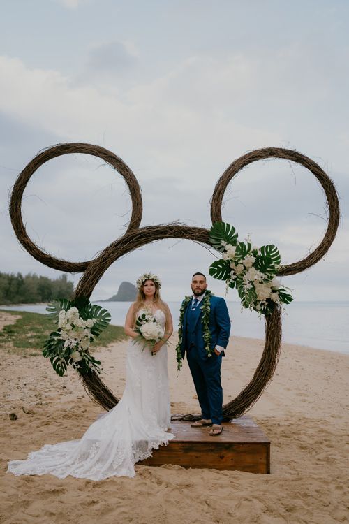 A Bride And Groom Are Standing In Front Of A Mickey Mouse Arch On The Beach - Honolulu, HI - Bella Rosa Corporation