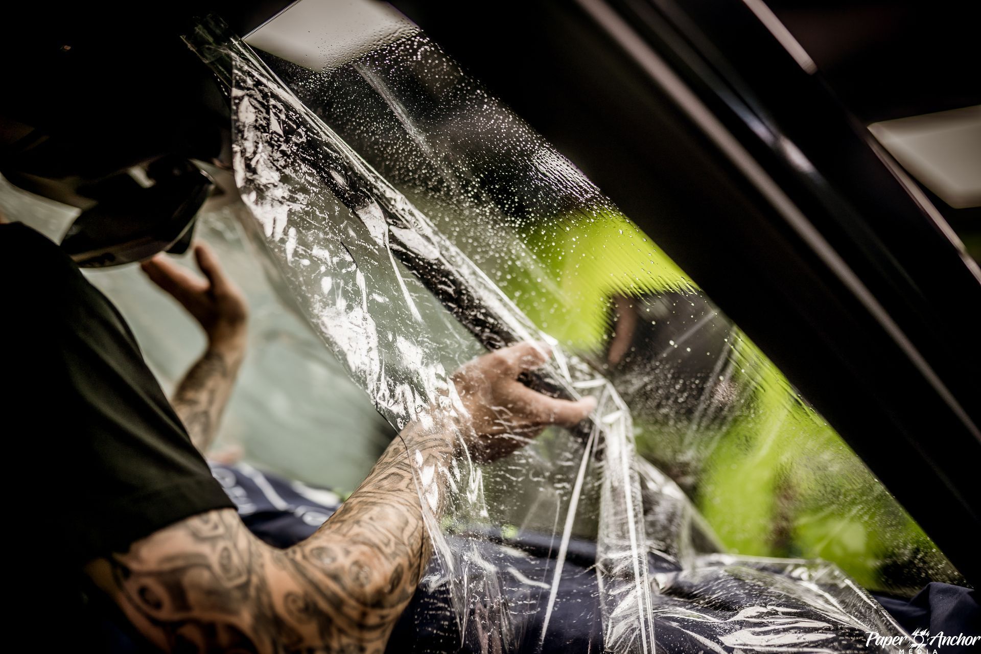 A person with tattoos applying tint to a car window. They are wearing a hat, inside a shop.