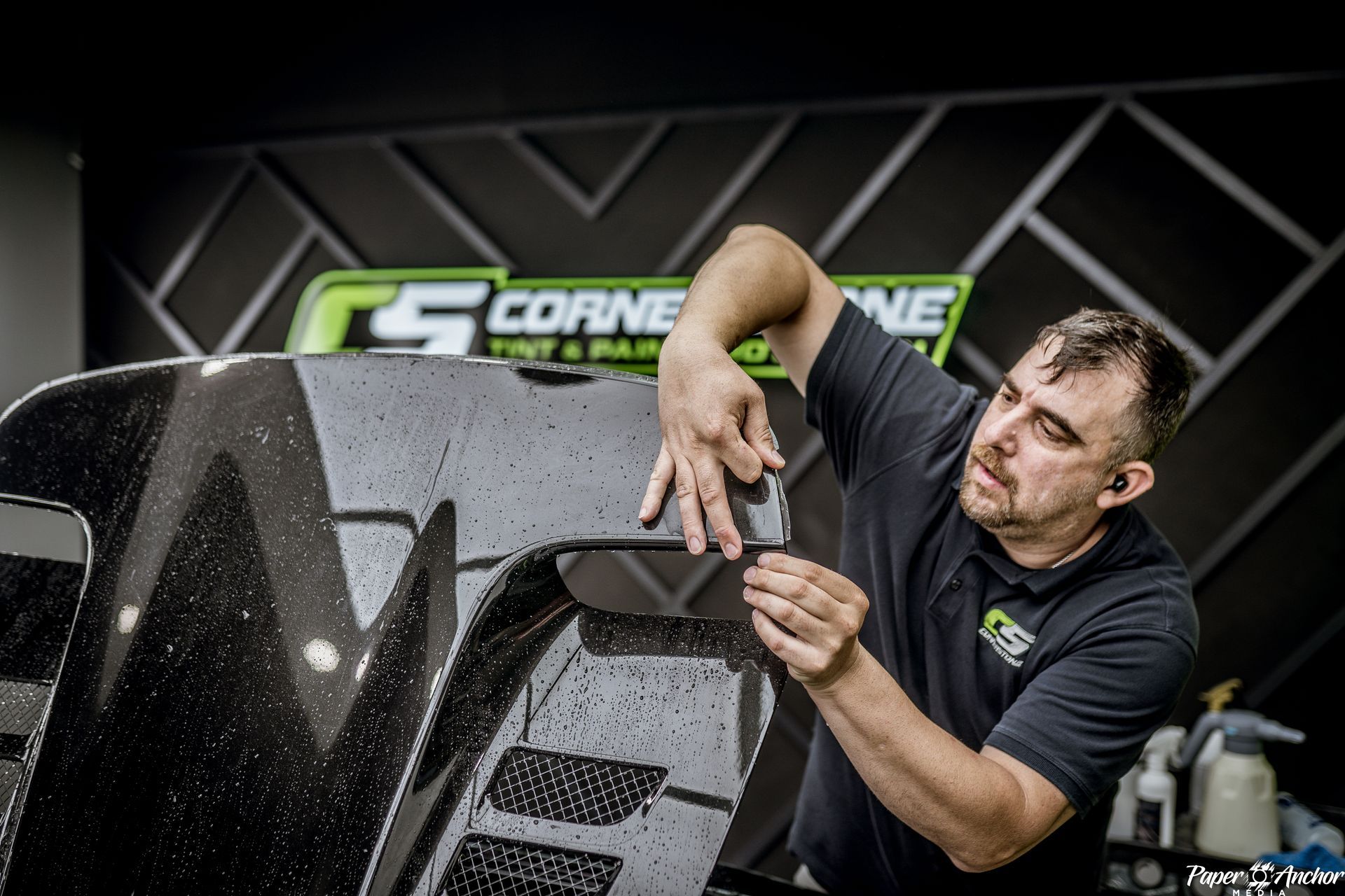 Man applies tint to a black car part, workshop setting, Cornerstone logo visible.