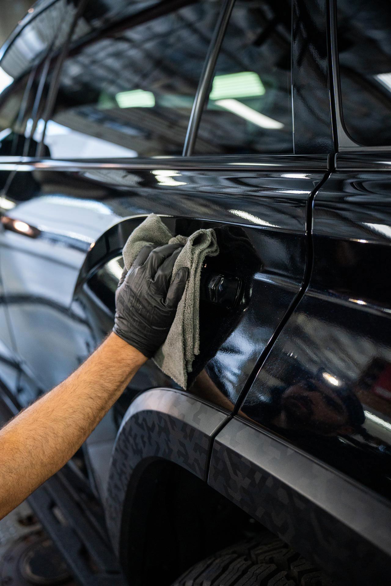 Gloved hand wiping a black car with a gray cloth. Focus on the car's fuel door and side panel.