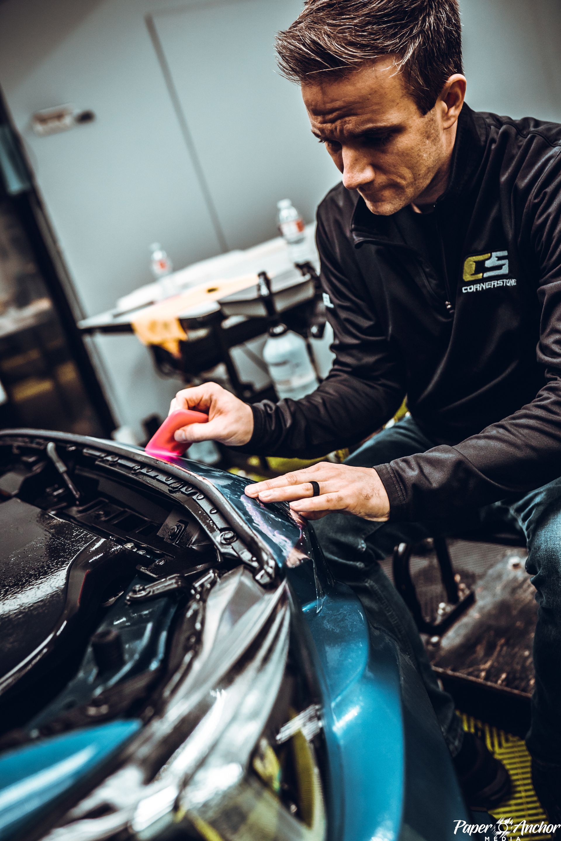 Man applying protective film to a teal car in a garage, using a squeegee.