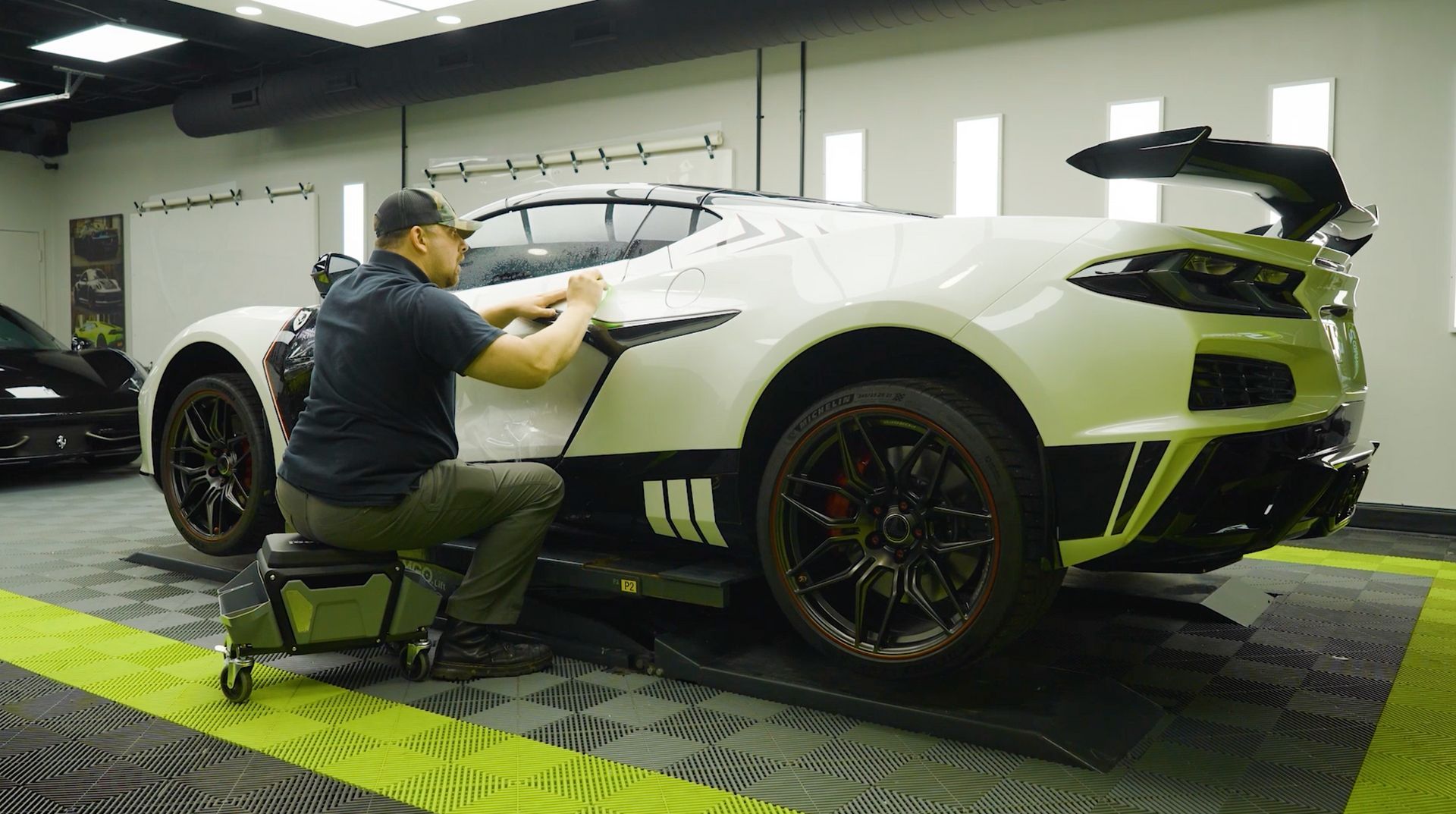 Man applies decal to white sports car in a garage; black wheels, spoiler.