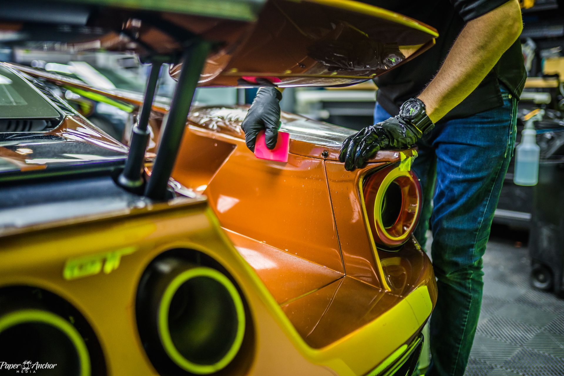 Person applying orange vinyl wrap to the rear of a sports car. Black gloves, blue jeans.