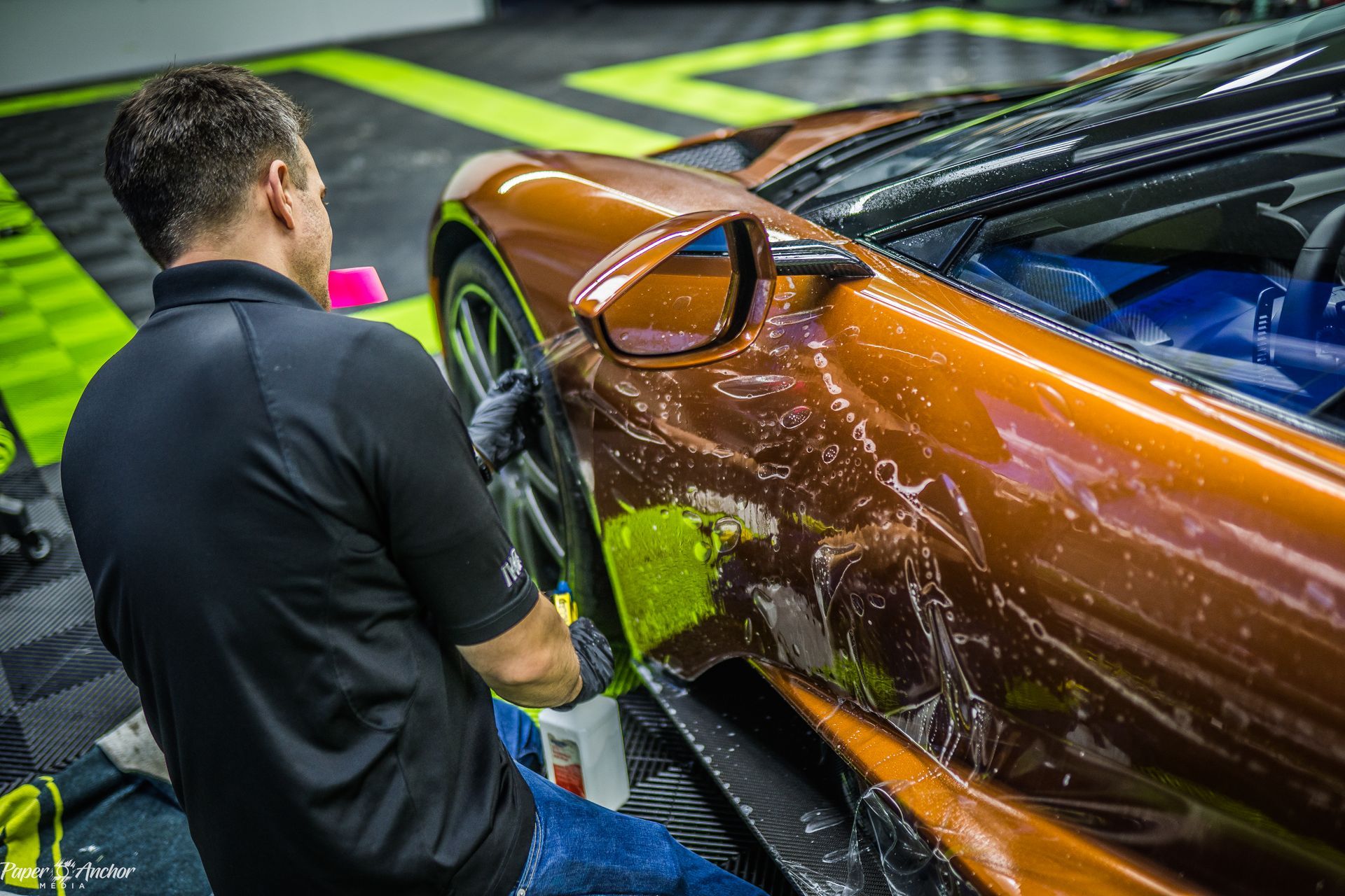 Man applying film to orange sports car in a garage with black and green flooring.