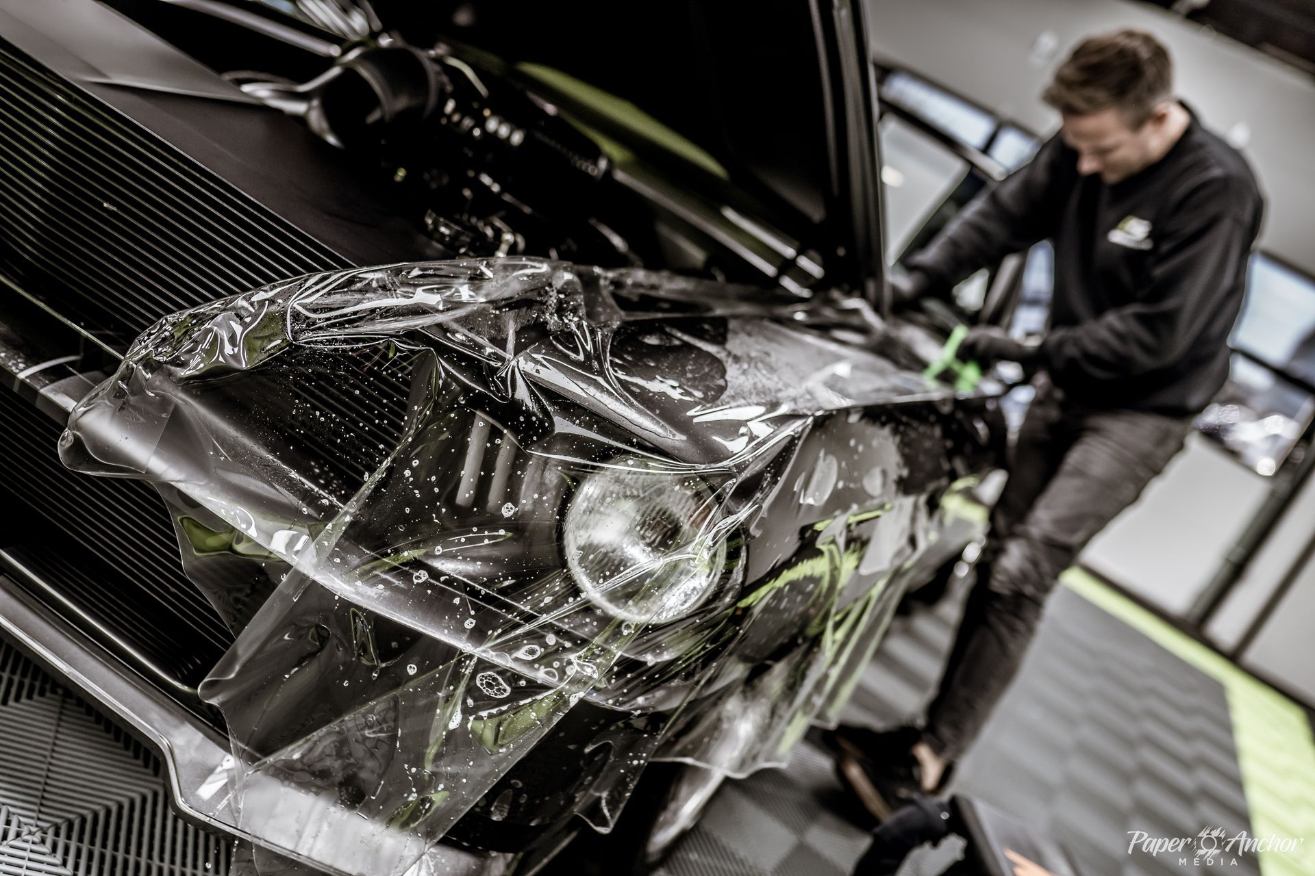 Man applying clear protective film to black car's headlight in a garage.